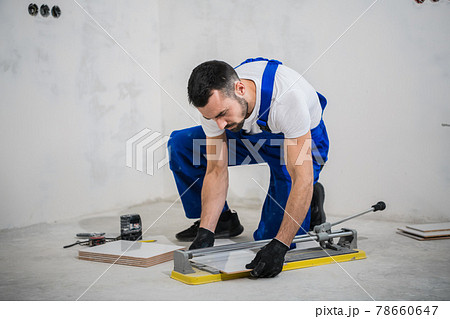 A worker in a blue overalls cuts a tile with a cutter A worker in a blue overalls cuts a tile with a cutter 78660647
