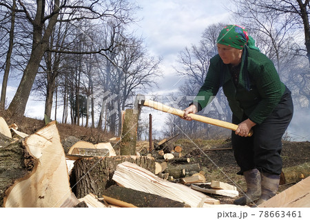 An old woman chopping firewood for the winter, also removes her yard near the house, on the street early spring. 78663461