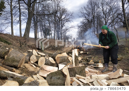 An old woman of a tavern shakes a firewood with an ax in the yard, prepares them for the winter. An old woman of a tavern shakes a firewood with an ax in the yard, prepares them for the winter. 78663470