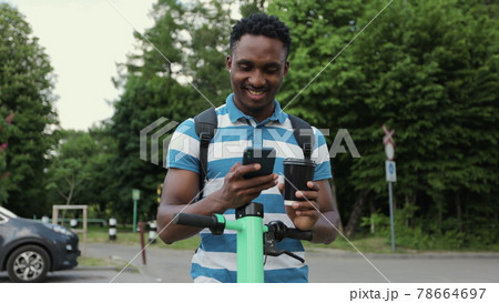 Afro man use smartphone stand with electric scooter and sipping hot drink to go. Man uses an app on his phone to rent an electric scooter. Ecological transportation. Urban outdoors. 78664697