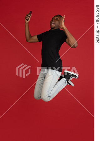 always on mobile. Full length of african young man taking phone while jumping against red studio background. Mobile, motion, movement, business concepts 78665598