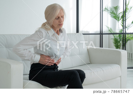 An elderly woman suffers from abdominal pain, a mature elderly adult woman feels abdominal pain while sitting on the couch An elderly woman suffers from abdominal pain, a mature elderly adult woman feels abdominal pain while sitting on the couch 78665747