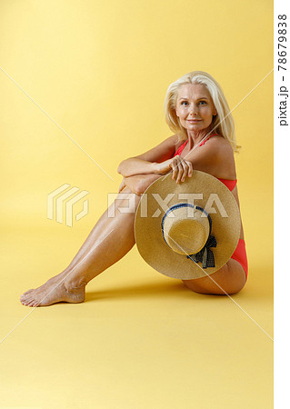 Graceful mature blonde woman in red swimsuit smiling at camera, holding straw hat while sitting on studio floor, posing isolated over yellow background 78679838