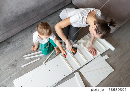 Cute adorable caucasian toddler boy kid sit on floor and help mom assembling furniture shelf with power screwdriver tool. Young adult mum with funny little child enjoy playing at home indoors Cute adorable caucasian toddler boy kid sit on floor and help mom assembling furniture shelf with power screwdriver tool. Young adult mum with funny little child enjoy playing at home indoors 78680395