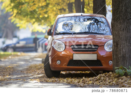Front view of small orange mini car parked in quiet yard on sunny autumn day on blurred buildings and big old trees golden foliage bokeh background. Transportation, parking problems concept. 78680871