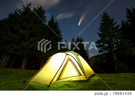 Illuminated tourist tent on green clearing with distant mountain and starry night sky with C/2020 F3 (NEOWISE) comet with light tail on it. 78681436