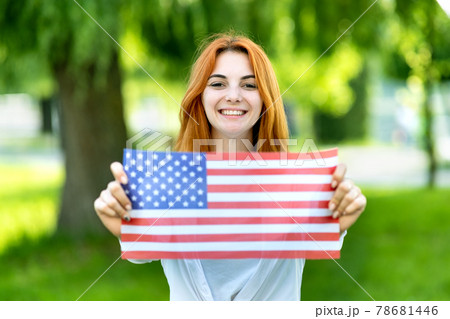 Portrait of happy young red haired woman holding USA national flag in her hands standing outdoors in summer park. Positive girl celebrating United States independence day. International day of Portrait of happy young red haired woman holding USA national flag in her hands standing outdoors in summer park. Positive girl celebrating United States independence day. International day of 78681446