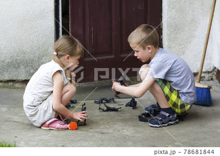 Two cute children blond pretty preschool girl and handsome boy playing outdoors with plastic toys on warm summer day on background of old house. Careless happy childhood and friendship concept. Two cute children blond pretty preschool girl and handsome boy playing outdoors with plastic toys on warm summer day on background of old house. Careless happy childhood and friendship concept. 78681844