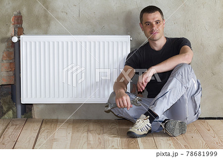 Young handsome plumber sitting on floor with wrench in hand near successfully installed heating radiator in empty room of a newly built apartment or house. Construction, maintenance and repair concept Young handsome plumber sitting on floor with wrench in hand near successfully installed heating radiator in empty room of a newly built apartment or house. Construction, maintenance and repair concept 78681999