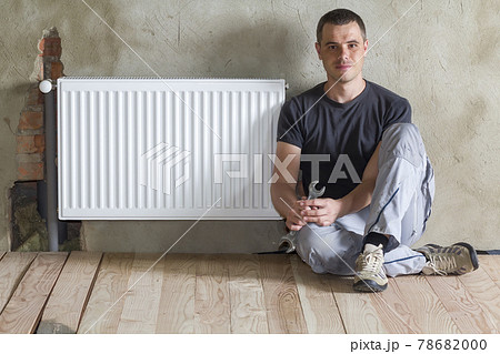 Young handsome plumber sitting on floor with wrench in hand near successfully installed heating radiator in empty room of a newly built apartment or house. Construction, maintenance and repair concept Young handsome plumber sitting on floor with wrench in hand near successfully installed heating radiator in empty room of a newly built apartment or house. Construction, maintenance and repair concept 78682000