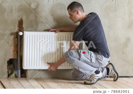 Young handsome professional plumber worker installing heating radiator in an empty room of a newly built apartment or house. Construction, maintenance and repair concept. Young handsome professional plumber worker installing heating radiator in an empty room of a newly built apartment or house. Construction, maintenance and repair concept. 78682001