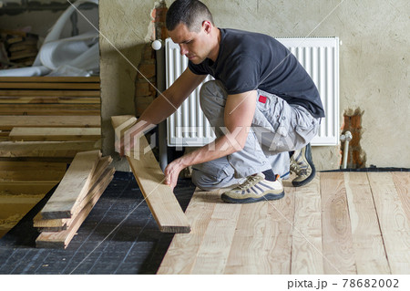 Young handsome professional carpenter installing natural wooden planks on wooden frame floor in empty unfinished room under reconstruction. Improvement, renovation and carpentry concept 78682002
