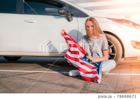 Young millennial blonde woman sitting with american flag next to car. Flag of the united states in her hands. July 4th Independence Day. USA national holiday. Freedom and memorial concept Young millennial blonde woman sitting with american flag next to car. Flag of the united states in her hands. July 4th Independence Day. USA national holiday. Freedom and memorial concept 78688671