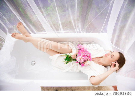 Beautiful girl in a white bathrobe with a bouquet of pink peonies near the bath. Beautiful girl in a white bathrobe with a bouquet of pink peonies near the bath. 78693336
