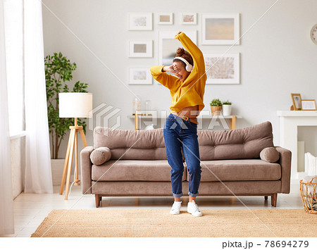 Young playful african american woman in headphones jumping and dancing in living room at home 78694279