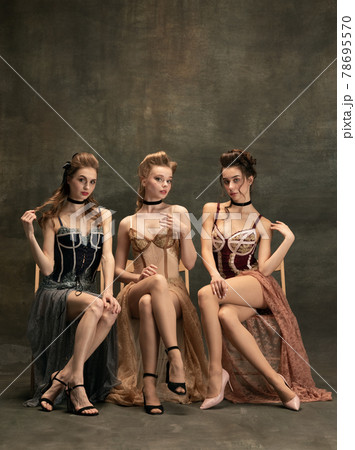 Pretty women in vintage 20s dresses. Young girls in art action sitting together and posing on wall background. Pretty women in vintage 20s dresses. Young girls in art action sitting together and posing on wall background. 78695570