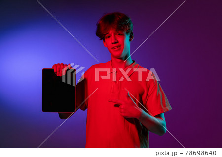 Holding tablet, crazy excited. Caucasian young man's portrait on black studio background in neon light. Beautiful male model in white shirt. 78698640