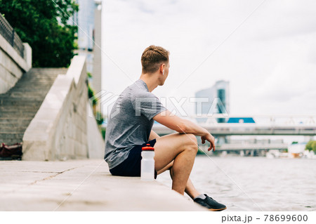 Tired Young man runner sitting and relaxing after sport training. Holding water bottle while doing workout in summer city street. 78699600