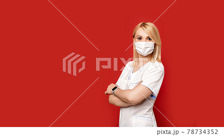 Confident female doctor wearing protective face mask standing on red background arms crossed. Young woman intern in medical clothes posing. 78734352
