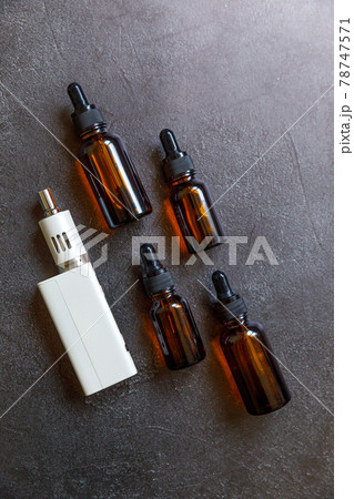 Vaping device e-cigarette electronic cigarette and liquid bottles on dark black stone shale background. Vape device for alternative smoking. Vaping shop concept. Gadget for vaper. Vaping accessories. 78747571