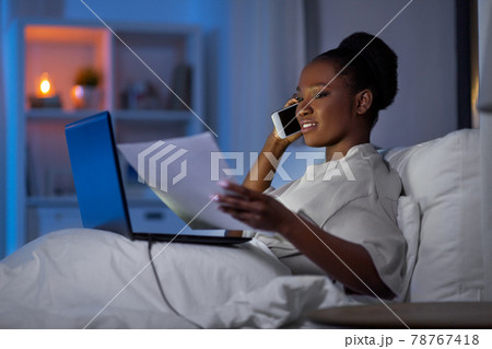 woman with laptop calling on smartphone in bed 78767418
