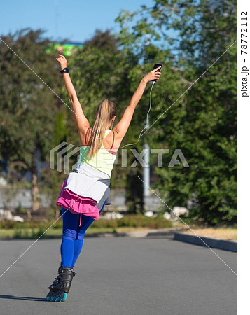 Back view of a young woman while rollerblading. She listens to music through headphones from her smartphone and enjoys life. 78772112