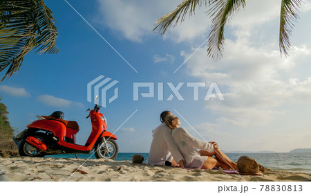 Scooter road trip. Lovely couple on red motorbike in white clothes on sand beach. People walking near the tropical palm trees, sea. Motorcycle rent. 78830813