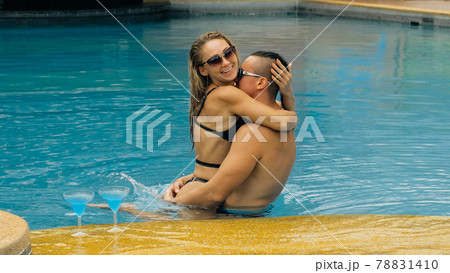 The loving couple hugs and kisses, drinking blue cocktail alcohol liquor in swimming pool at hotel outdoor. Portrait of caucasian man and woman. The loving couple hugs and kisses, drinking blue cocktail alcohol liquor in swimming pool at hotel outdoor. Portrait of caucasian man and woman. 78831410