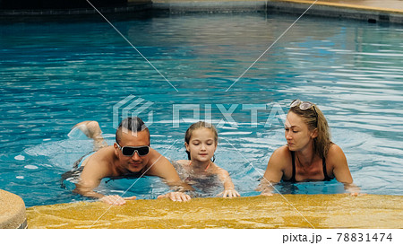 The mother and father with little daughter have fun in the pool. Mom and dad plays with the child. The family enjoy summer vacation in a swimming. 78831474