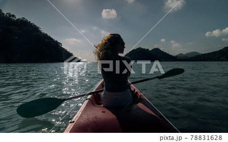 Silhouette of woman tourist raising hands while sailing in canoe along sea bay water to distant hilly island backside view. Silhouette of woman tourist raising hands while sailing in canoe along sea bay water to distant hilly island backside view. 78831628