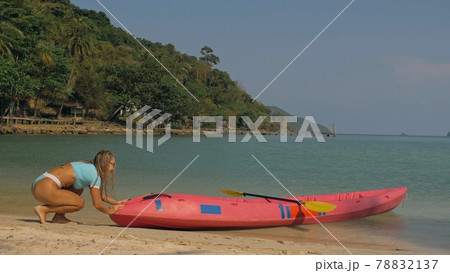 Pretty young sportswoman with sunglasses and swimsuit holds paddle posing near pink plastic canoe on sand beach at resort. Pretty young sportswoman with sunglasses and swimsuit holds paddle posing near pink plastic canoe on sand beach at resort. 78832137