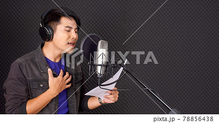 Young asian man with headphones sing while holding lyrics in his hand in front of black soundproofing walls. Musicians producing music in professional recording studio. Young asian man with headphones sing while holding lyrics in his hand in front of black soundproofing walls. Musicians producing music in professional recording studio. 78856438