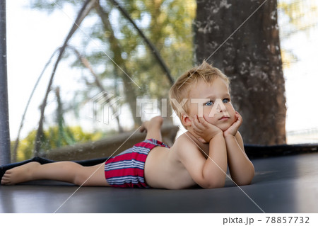 Portrait of cute bored little caucasian funny sad blond toddler boy lying inside big black trampoline at home backyard playground area outdoors warm summer sunny day. Children street sport activity Portrait of cute bored little caucasian funny sad blond toddler boy lying inside big black trampoline at home backyard playground area outdoors warm summer sunny day. Children street sport activity 78857732