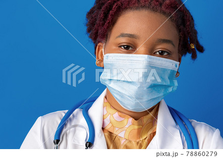 Portrait of african female doctor in lab coat with face mask and stethoscope against blue background Portrait of african female doctor in lab coat with face mask and stethoscope against blue background 78860279