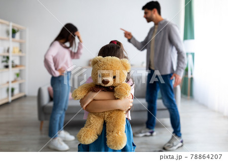 Children and family conflicts concept. Little girl cuddling teddy bear standing against arguing parents Children and family conflicts concept. Little girl cuddling teddy bear standing against arguing parents 78864207