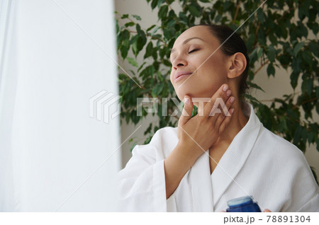 Young African American woman in white bathrobe caring of her skin, applying face moisturizer , tenderly massaging her neck 78891304