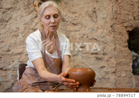 Elderly female potter in an apron makes a pot in a pottery workshop Elderly female potter in an apron makes a pot in a pottery workshop 78894517
