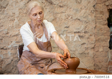 Elderly female potter in an apron makes a pot in a pottery workshop Elderly female potter in an apron makes a pot in a pottery workshop 78894518