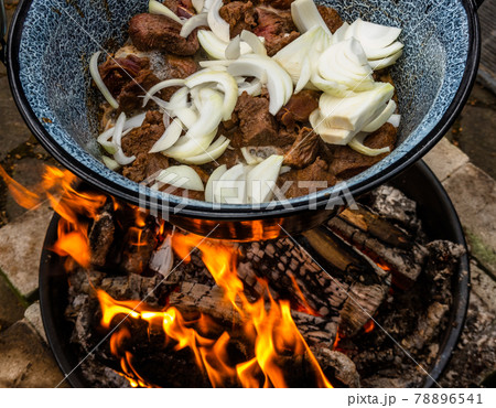 White onions and roasted pieces of beef hang in a pot over an open wood fire for the preparation of kettle goulash, camping 78896541