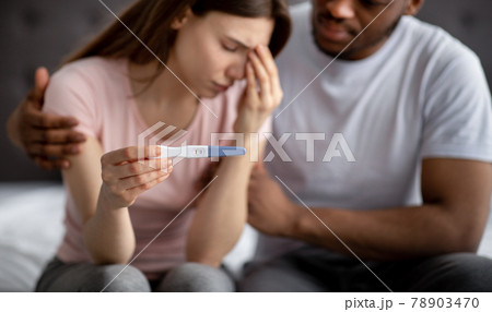 Upset young woman showing negative pregnancy test, her black husband supporting her in bedroom, selective focus 78903470