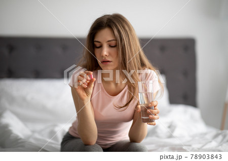 Millennial lady with glass of water taking pill on bed, treating her illness at home 78903843