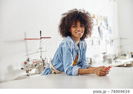 African-American seamstress with smartphone rests leaning onto cutting table in workshop 78909639