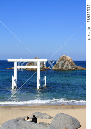 波打ち際の岩に座り桜井二見ヶ浦の白い鳥居と夫婦岩を眺めるカップル 78925537