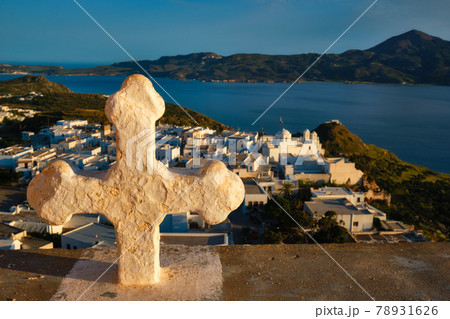 Christian cross and Plaka village on Milos island over red geranium flowers on sunset in Greece 78931626