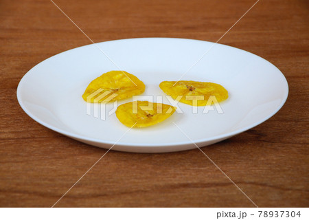Dried and fried banana slices place on white plate isolated on wood texture background. This is local Malaysian traditional junk food call Kerepek Pisang. Dried and fried banana slices place on white plate isolated on wood texture background. This is local Malaysian traditional junk food call Kerepek Pisang. 78937304