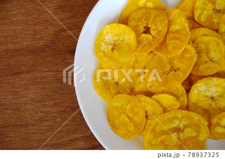 Dried and fried banana slices place on white plate isolated on wood texture background. This is local Malaysian traditional junk food call Kerepek Pisang. Dried and fried banana slices place on white plate isolated on wood texture background. This is local Malaysian traditional junk food call Kerepek Pisang. 78937325