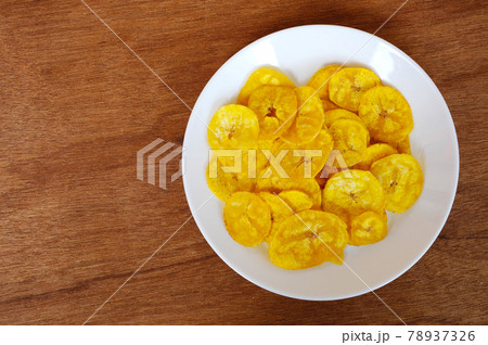 Dried and fried banana slices place on white plate isolated on wood texture background. This is local Malaysian traditional junk food call Kerepek Pisang. Dried and fried banana slices place on white plate isolated on wood texture background. This is local Malaysian traditional junk food call Kerepek Pisang. 78937326
