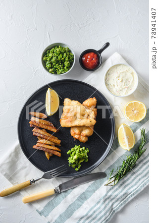 British Traditional Fish and chips with minty mashed peas detail and a slice of lemon. on black round plate over white lintn and stone surface top view 78941407