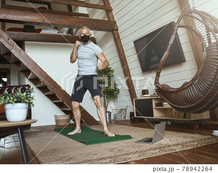 Covid 19 outbreak. Active middle aged man in face protective mask standing in boxing stance in the living room at home, watching online workout on laptop Covid 19 outbreak. Active middle aged man in face protective mask standing in boxing stance in the living room at home, watching online workout on laptop 78942264