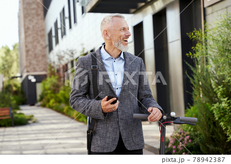 Joyful mature businessman holding smartphone, looking aside while walking with electric scooter outdoors on a daytime Joyful mature businessman holding smartphone, looking aside while walking with electric scooter outdoors on a daytime 78942387